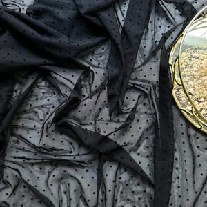 May include: Black sheer fabric with small, evenly spaced polka dots. The fabric is draped and folded, creating shadows and texture. A gold-rimmed decorative tray is visible in the upper right corner, adding a touch of elegance to the scene.