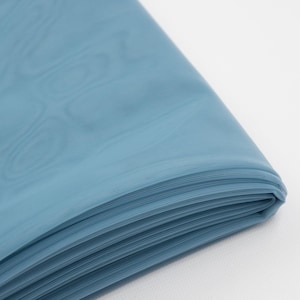 May include: A folded piece of dusty blue fabric with a fine, semi-sheer texture. The material is layered, showing multiple crisp folds along the edge, with a subtle, smooth surface finish that catches the light across its surface.