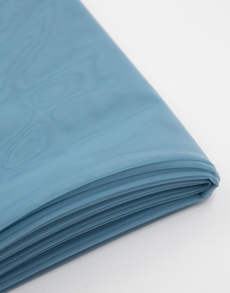 May include: A folded piece of dusty blue fabric with a fine, semi-sheer texture. The material is layered, showing multiple crisp folds along the edge, with a subtle, smooth surface finish that catches the light across its surface.