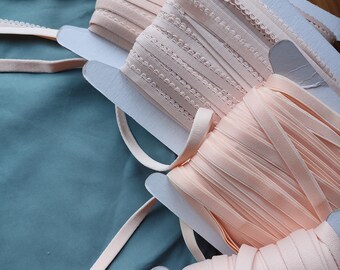 Può includere: Quattro bobine di passamaneria elastica rosa con diversi motivi e larghezze. La passamaneria è su bobine di cartone bianco.