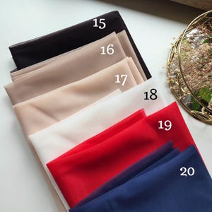 May include: A collection of six folded sheer fabric swatches in black, beige, light tan, white, red, and navy blue. Each piece of fabric is labelled with a white number from 15 to 20, indicating distinct colour options for the material.