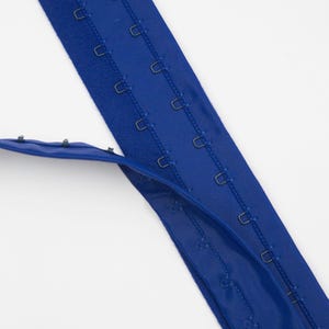 May include: A royal blue bra extender featuring a long fabric strip with multiple rows of metal hook-and-eye closures. The design includes a vertical column of eyelets on one side and a corresponding row of metal hooks on the other side.