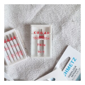 Puede incluir: Primer plano de un paquete de agujas para m&aacute;quina de coser Organ. El paquete es blanco con texto rojo que dice "ORGAN 130/705H TWIN 90/4".