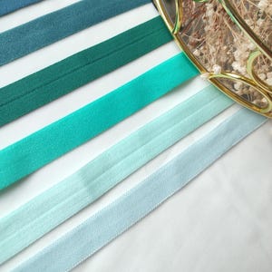 May include: A set of seven satin ribbons in shades of blue and green. The ribbons are arranged in a gradient from dark blue to light blue, with a green ribbon in the middle. The ribbons are all about 1 inch wide.