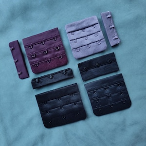 Può includere: Assortimento di prolunghe per spalline del reggiseno in vari colori e dimensioni. Le prolunghe sono realizzate in tessuto nero, viola e grigio con ganci e occhielli in metallo.