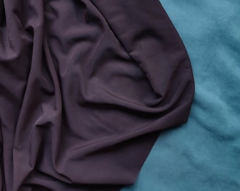 May include: A close-up shot of draped, dark purple fabric against a teal background. The fabric is soft and flowing, with visible folds and shadows. The teal background has a velvety texture, creating a contrast with the smooth purple material.