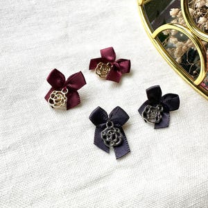 Könnte beinhalten: Vier kleine Schleifen aus bordeauxfarbenem und dunkelgrauem Band, jeweils mit einem Rosenanhänger aus Metall. Die Schleifen sind auf einer weißen, strukturierten Oberfläche angeordnet. Ein goldgerahmter Spiegel befindet sich im Hintergrund.