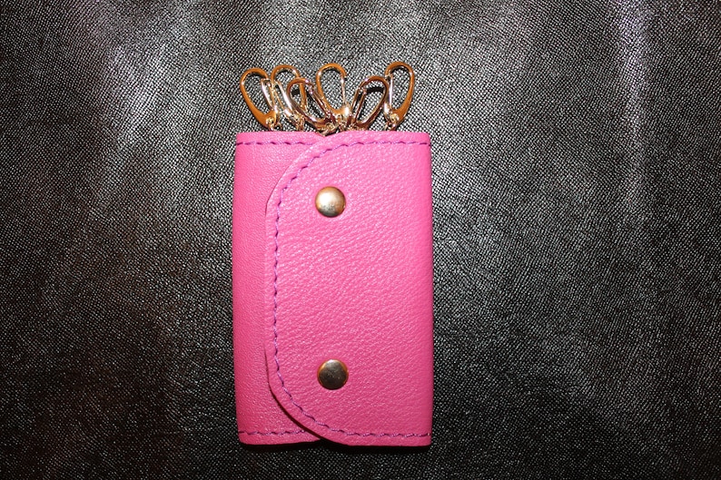 May include: A pink leather key holder with gold key rings. The key holder has a flap with two gold snaps.