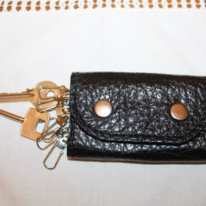 May include: A black leather key holder with two silver snaps and a flap closure. The key holder is filled with a set of keys and key rings.