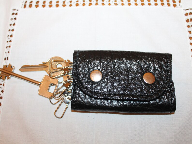 May include: A black leather key holder with two silver snaps and a flap closure. The key holder is filled with a set of keys and key rings.