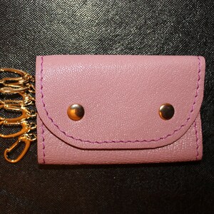 May include: A mauve leather key holder with gold-colored hardware. The key holder has a flap closure secured by two gold-colored snaps. The edges are stitched with purple thread, and a chain with multiple key rings is attached.