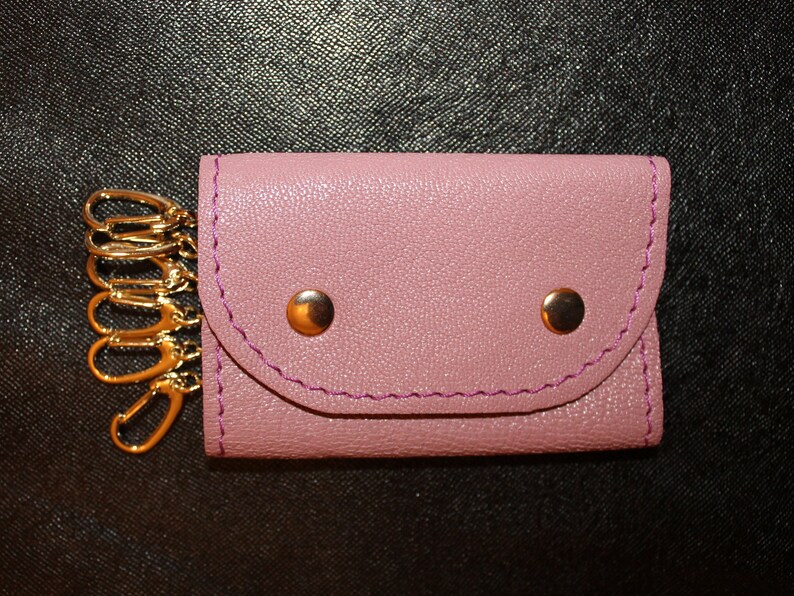 May include: A pink leather key holder with a gold key chain and two gold snaps. The key holder has a flap that folds over to secure the keys.