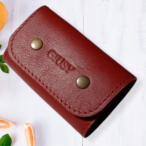 May include: A burgundy leather key holder with the word "GLUSY" embossed on the front. The holder has two silver snaps.