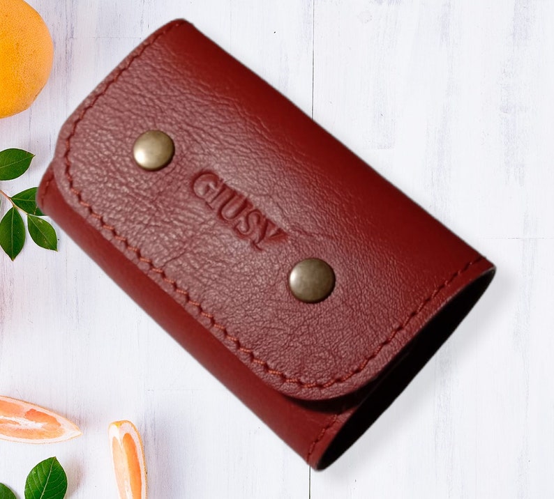 May include: A burgundy leather key holder with the word "GLUSY" embossed on the front. The holder has two silver snaps.