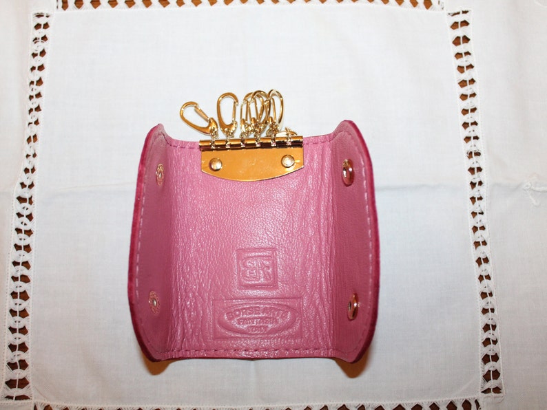May include: A pink leather key holder with a gold metal key ring and a small metal plate with the text "Genuine Leather" on it.
