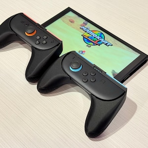 May include: A black games console with two detached controllers. One controller has an orange button, and the other has a blue button. The screen displays the "Mario Kart World" game.