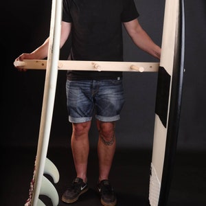 May include: A wooden surfboard rack with three surfboards hanging on it. The rack is made of natural wood and has a simple design. The surfboards are white with black stripes and fins.