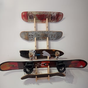 Pode incluir: Um suporte de parede de madeira com quatro skates e um snowboard. Os skates estão dispostos verticalmente, com o snowboard na parte inferior. Os skates têm vários designs, incluindo um design vermelho e preto, um design preto e branco e um design marrom e branco. O snowboard é vermelho, laranja e amarelo com fixações pretas.