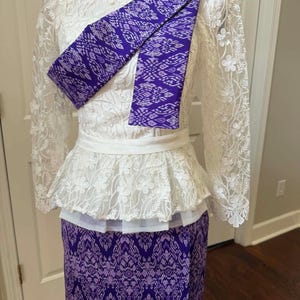 Pode incluir: Um conjunto de duas peças com um top branco de renda de manga comprida com cintura peplum e uma saia roxa com um padrão intrincado repetido. Uma faixa roxa a condizer com o mesmo padrão está drapeada diagonalmente sobre o corpete de renda.