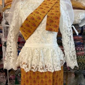 Puede incluir: Un vestido tailandés tradicional exhibido en un maniquí. El vestido presenta un corpiño de encaje blanco con mangas largas y una falda con volantes. Una faja dorada con un estampado floral rojo y verde está drapeada sobre los hombros. La falda es de color dorado a juego con un diseño floral similar.