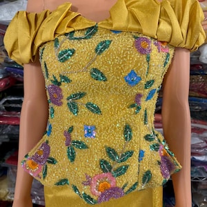 Puede incluir: Conjunto de top y falda amarillos con hombros descubiertos. El top presenta un corpiño ajustado con bordados florales y de hojas en lentejuelas moradas, rosas, verdes y azules. Las mangas son con volantes. La falda es amarilla lisa.