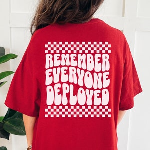 May include: Red t-shirt with a white checkered border and the text "Remember Everyone Deployed" in white, wavy letters.