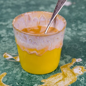 Könnte beinhalten: Eine gelbe Keramiktasse mit weißem Überzug und einem silbernen Löffel darin. Die Tasse ist mit einer braunen Flüssigkeit gefüllt, möglicherweise Kaffee oder Tee. Die Tasse steht auf einer grün-gelb gemusterten Oberfläche.