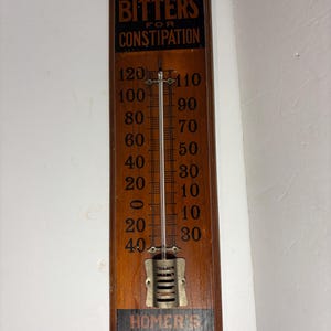 May include: Antique wooden thermometer with the text "LASH'S BITTERS FOR CONSTIPATION" in black and gold lettering. The thermometer has a scale from -40 to 120 degrees Fahrenheit. The bottom of the thermometer has the text "HOMER'S GINGER BRAND".