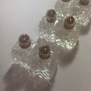 May include: A collection of clear, faceted glass salt and pepper shakers with gold-colored metal lids. The shakers are arranged in a row, casting shadows on a white surface. The design of the glass gives the shakers a vintage look.