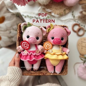 Puede incluir: Dos muñecas de cerdito de ganchillo rosa en una cesta de mimbre. Los cerdos tienen detalles, visten vestidos y sostienen piruletas. El texto "PDF PATTERN" está encima de la cesta. Las muñecas están hechas a mano.