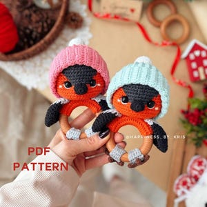 May include: Two handmade crochet bird toys, each with a wooden ring and wearing a hat. One bird has a pink hat, the other a light blue hat. The birds have orange bodies, black wings, and grey accents. The text "PDF PATTERN" is visible.