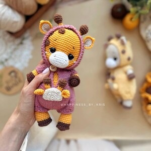 May include: A crocheted giraffe doll in a pink hooded jumpsuit. The giraffe features yellow and brown details on its ears, horns, and feet. The doll is held in a hand. The text "@HAPPINESS_BY_KRIS" is visible. A second giraffe doll is in the background.