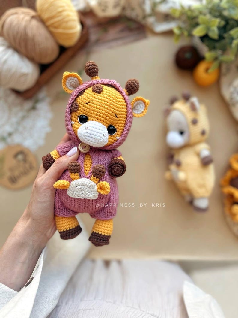May include: A crocheted giraffe doll in a pink hooded jumpsuit. The giraffe features yellow and brown details on its ears, horns, and feet. The doll is held in a hand. The text "@HAPPINESS_BY_KRIS" is visible. A second giraffe doll is in the background.