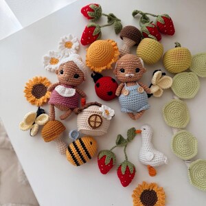 K&ouml;nnte beinhalten: Eine Sammlung geh&auml;kelter Objekte, darunter zwei kleine Puppen, ein Marienk&auml;fer, eine Biene, eine wei&szlig;e Ente, Pilze, Erdbeeren, Sonnenblumen, G&auml;nsebl&uuml;mchen und ein kleines Haus, gefertigt aus Garn in verschiedenen Farben wie Rot, Gelb, Blau und Gr&uuml;n.