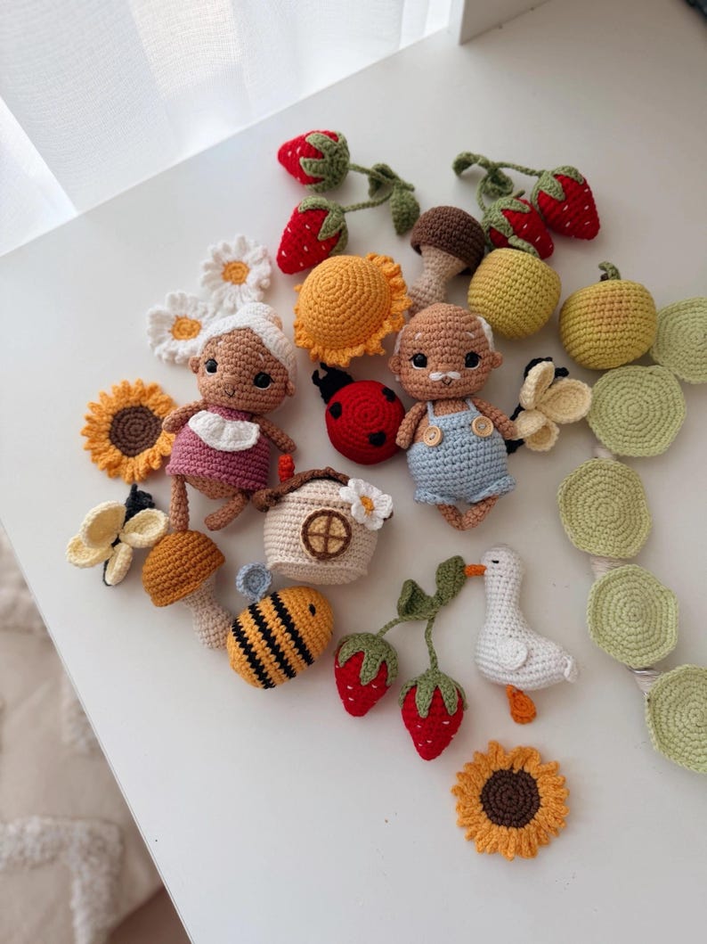 K&ouml;nnte beinhalten: Eine Sammlung geh&auml;kelter Objekte, darunter zwei kleine Puppen, ein Marienk&auml;fer, eine Biene, eine wei&szlig;e Ente, Pilze, Erdbeeren, Sonnenblumen, G&auml;nsebl&uuml;mchen und ein kleines Haus, gefertigt aus Garn in verschiedenen Farben wie Rot, Gelb, Blau und Gr&uuml;n.