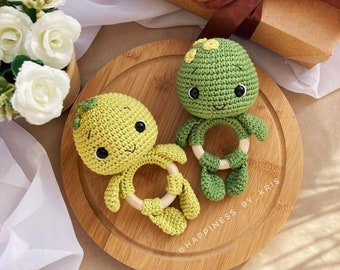 May include: Two handmade crochet turtle baby rattles in shades of green and yellow. Each turtle has a wooden ring and black safety eyes. The rattles are displayed on a wooden tray, with a gift box and flowers in the background.