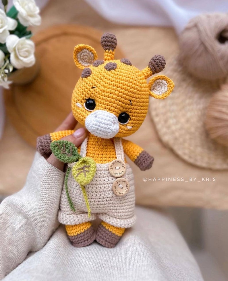 May include: A handmade crochet giraffe toy in yellow, brown, and cream colors. The giraffe has a white muzzle, black eyes, and wears cream-colored overalls with wooden buttons. It holds a green leaf. The text "@HAPPINESS_BY_KRIS" is visible.