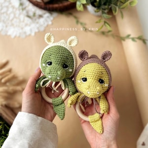 Pode incluir: Duas chocalhos de bebê feitos à mão em crochê, um verde e outro amarelo, cada um em forma de crocodilo com capuz e anel de madeira. Os chocalhos são segurados nas mãos contra um fundo neutro, com o texto "@HAPPINESS_BY_KRIS" visível.