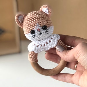 May include: A crocheted cat rattle toy with a wooden ring. The cat is pink and white with black eyes and a white collar.