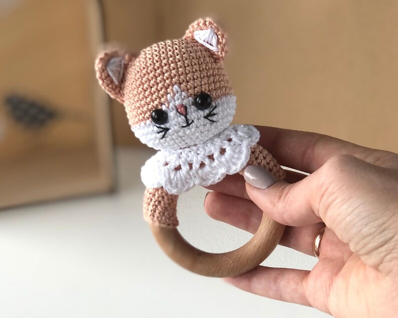 May include: A crocheted cat rattle toy with a wooden ring. The cat is pink and white with black eyes and a white collar.