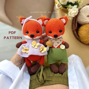 May include: Two crocheted fox amigurumi toys, one wearing a purple dress with yellow flowers and the other wearing a green vest and brown pants. The toys are sitting on a person's lap. The text "PDF PATTERN" is visible in the image.