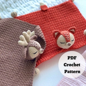 Peut inclure: Deux couvertures pour b&eacute;b&eacute; en crochet avec des applications d'animaux. Une couverture est marron avec une application de cerf et l'autre est orange avec une application de renard. Les couvertures sont dispos&eacute;es sur une surface blanche avec un tissu blanc et rose en arri&egrave;re-plan. Le texte "PDF Crochet Pattern" est visible sur l'image.