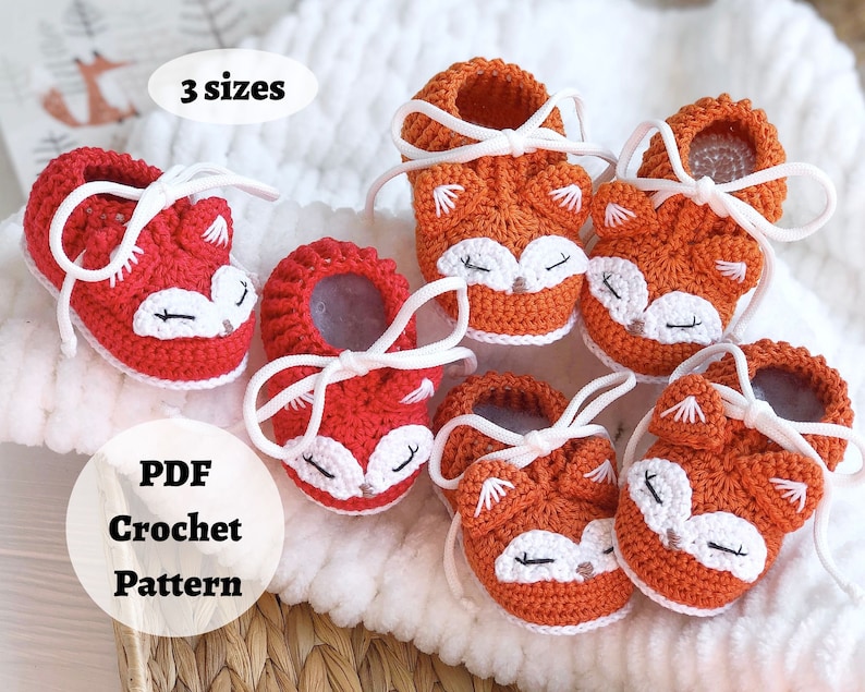 May include: Crochet pattern for fox baby booties in red and orange with white laces. The pattern includes three sizes. The text "PDF Crochet Pattern" is on a white circle.
