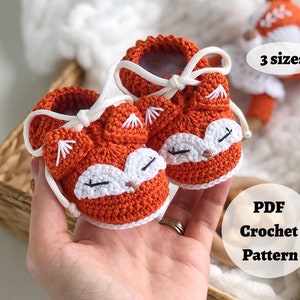 May include: A pair of orange crocheted baby booties with white fox faces and white laces. The booties are held in a hand. The text "3 sizes" and "PDF Crochet Pattern" is visible in the image.