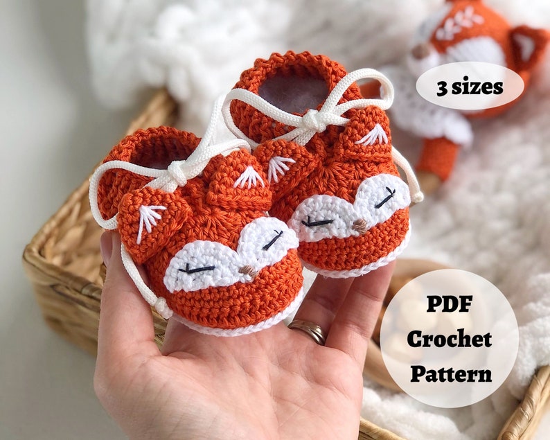 May include: A pair of orange crocheted baby booties with white fox faces and white laces. The booties are held in a hand. The text "3 sizes" and "PDF Crochet Pattern" is visible in the image.