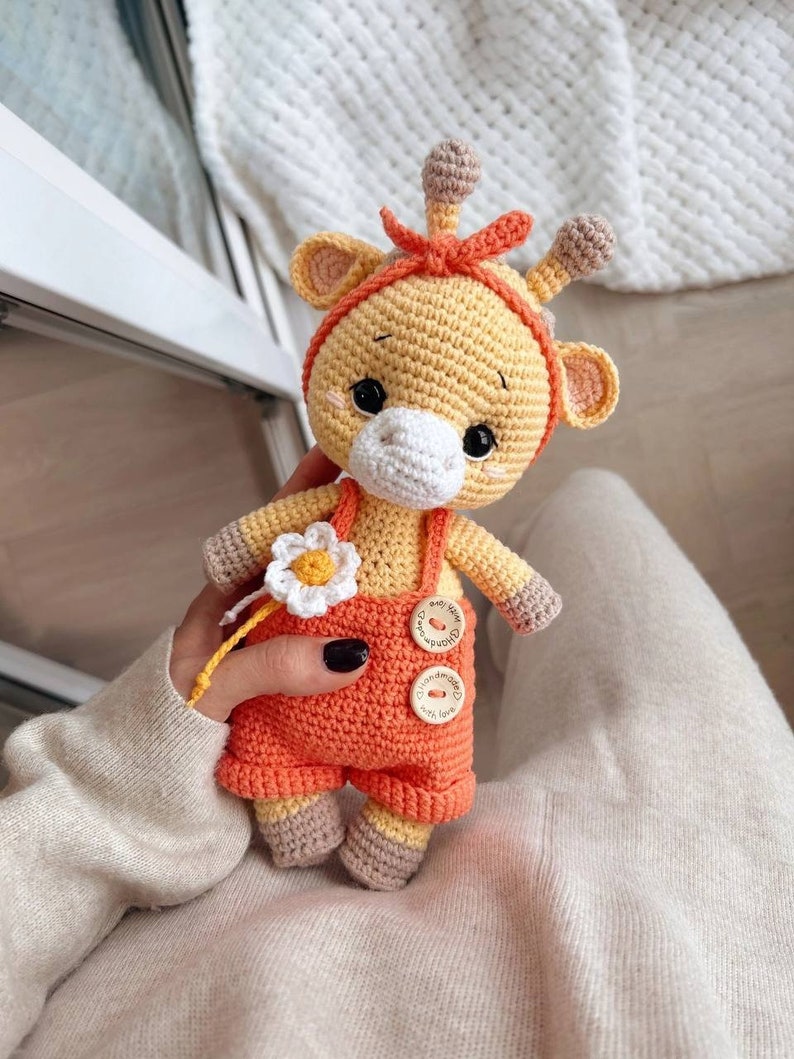 May include: A handmade crochet giraffe doll in shades of yellow, orange, and brown. The giraffe wears orange overalls with button details and a headband. It holds a white daisy with a yellow center. The doll is approximately 6 inches tall.
