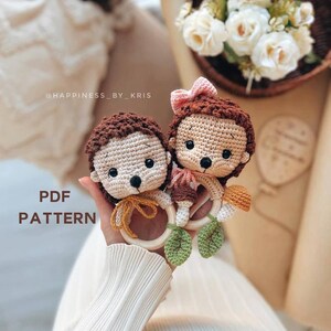 Puede incluir: Dos juguetes de erizo de mar hechos a crochet sosteniendo anillos de dentición de madera con hojas verdes de crochet. Los erizos son marrones y beige con ojos negros y narices marrones. El texto "PDF PATTERN" está escrito en letras marrones sobre un fondo blanco.