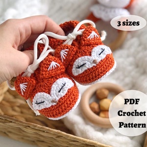 May include: A pair of orange and white crocheted baby booties shaped like foxes. The booties have white laces and are held in a hand. The text "3 sizes" and "PDF Crochet Pattern" is visible in the image.