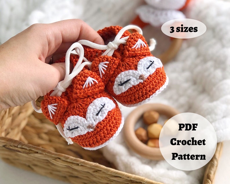 May include: A pair of orange and white crocheted baby booties shaped like foxes. The booties have white laces and are held in a hand. The text "3 sizes" and "PDF Crochet Pattern" is visible in the image.
