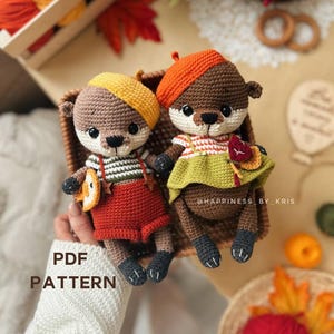 May include: Two crocheted otter dolls, each wearing a hat and outfit. One otter wears a yellow and brown hat, red shorts, and a striped shirt. The other otter wears an orange hat and a green and orange dress. The text "PDF PATTERN" is visible.